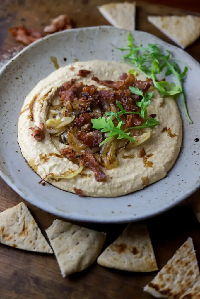 This bacon and caramelized onion hummus and pita bread appetizer. Yum. A different twist on your typical hummus flavors. It's a simple, homemade hummus topped with bacon, sweet, caramelized onion, and a drizzle of balsamic vinegar. This dip is so good I could make a meal out of it.